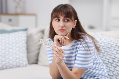 Photo of Upset woman with blister of contraceptive pills on sofa at home