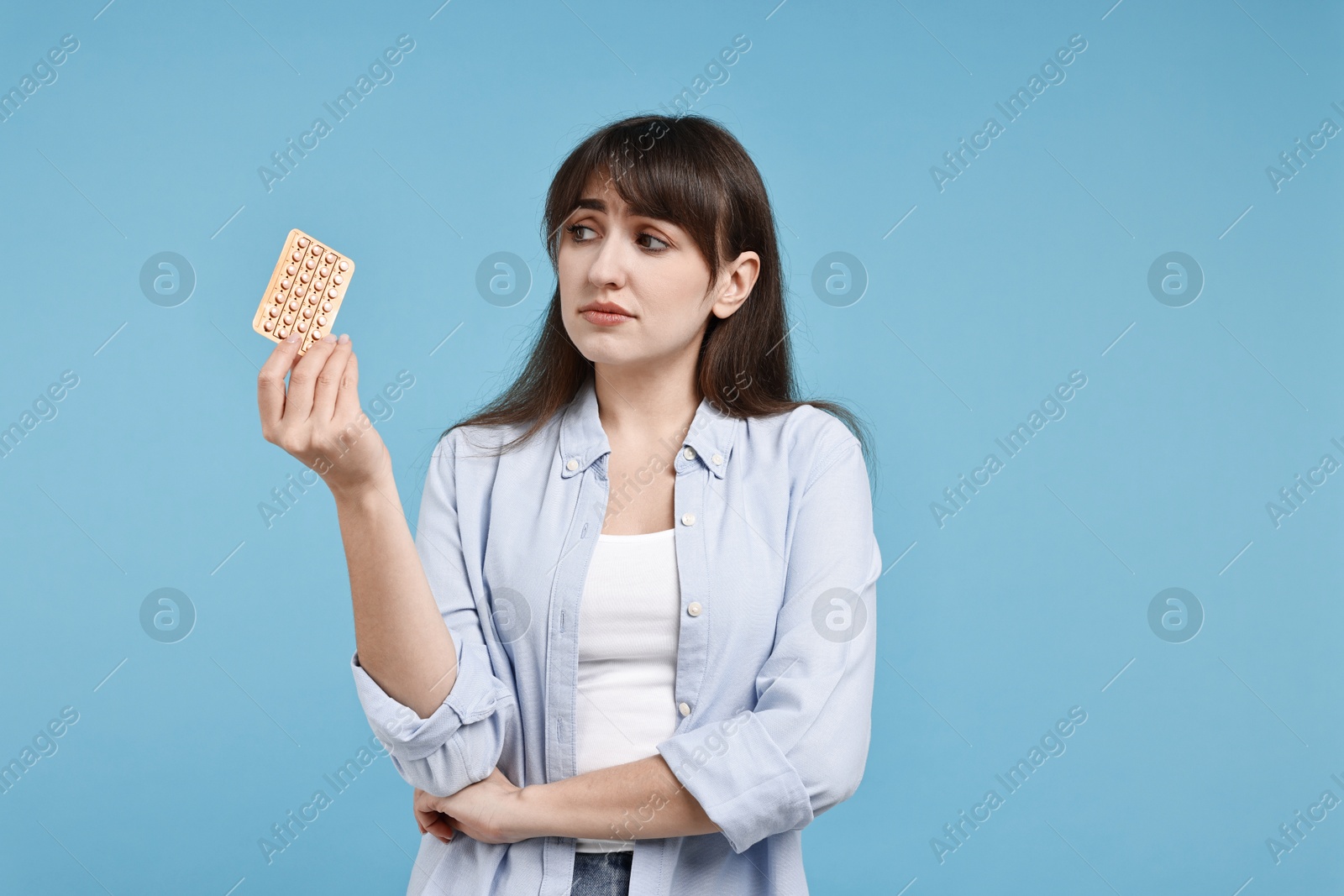 Photo of Sad woman with blister of contraceptive pills on light blue background