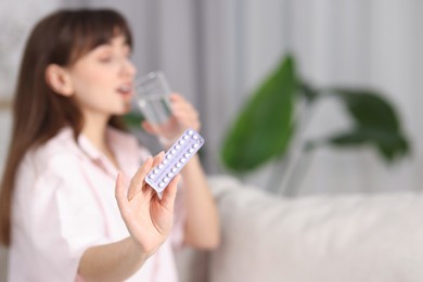 Photo of Woman with blister of contraceptive pills drinking water indoors, selective focus. Space for text