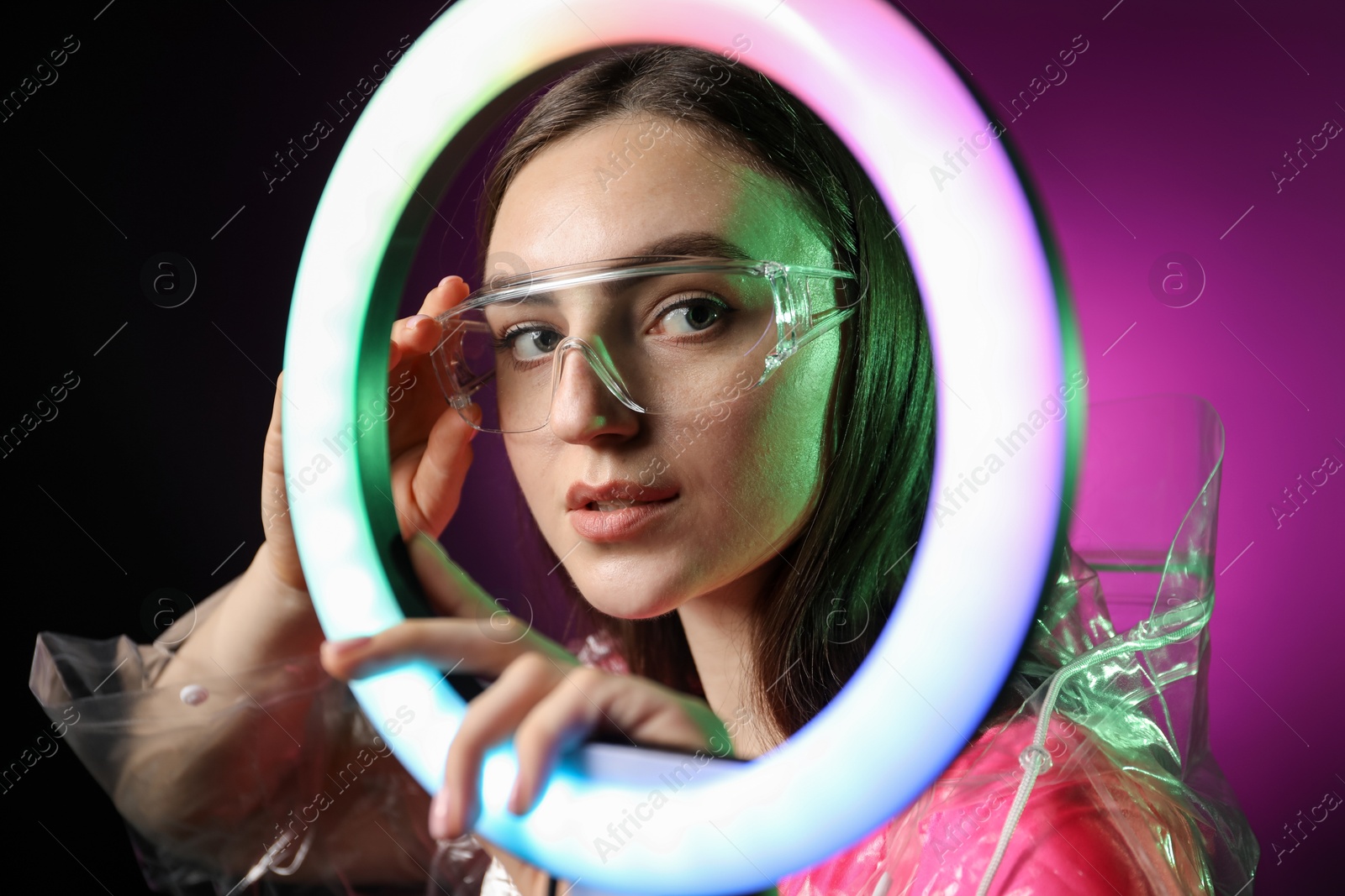 Photo of Stylish woman wearing transparent coat and glasses on dark purple background, view through ring lamp