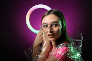 Photo of Stylish woman with ring lamp wearing transparent coat and glasses on dark purple background, space for text