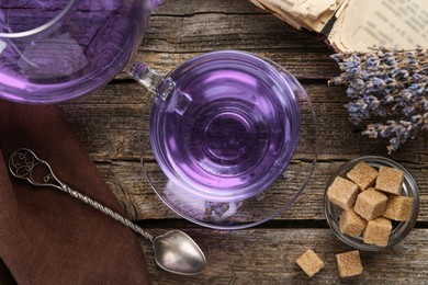 Photo of Aromatic lavender tea in glass cup, spoon, brown sugar, teapot, book and bunch of dry flowers on wooden table, flat lay