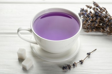 Photo of Aromatic lavender tea in cup, sugar cubes and dry flowers on white wooden table, closeup