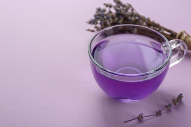 Photo of Aromatic lavender tea in glass cup and bunch of dry flowers on lilac background, closeup. Space for text