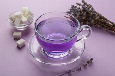 Photo of Aromatic lavender tea in glass cup, sugar cubes and dry flowers on lilac background, closeup