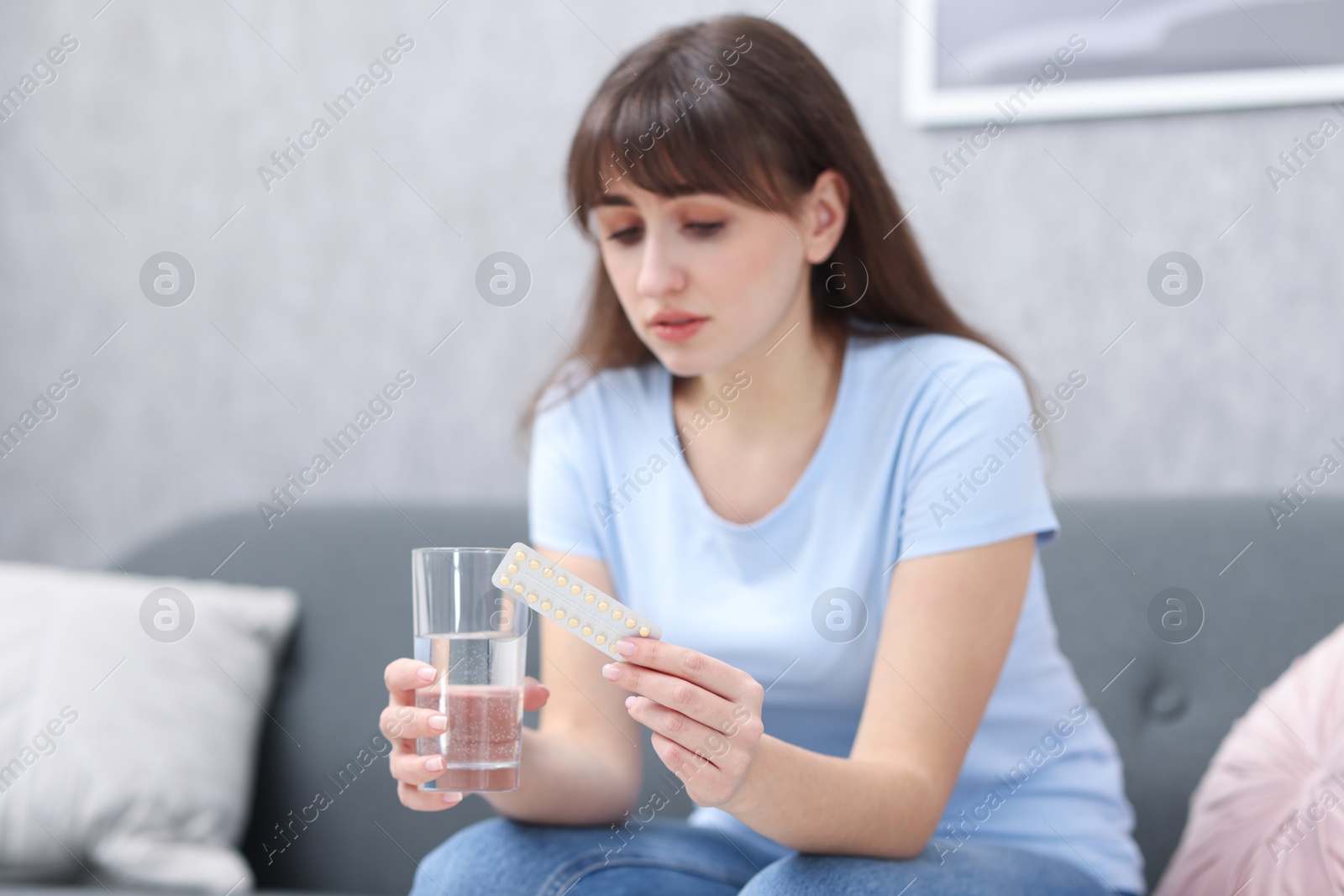 Photo of Sad woman with contraceptive pills and glass of water on sofa indoors