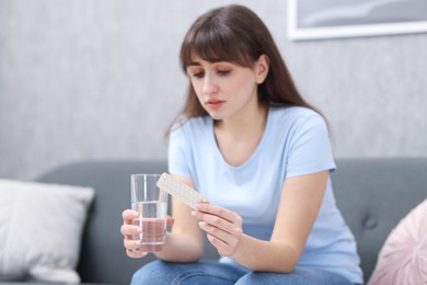 Photo of Sad woman with contraceptive pills and glass of water on sofa indoors