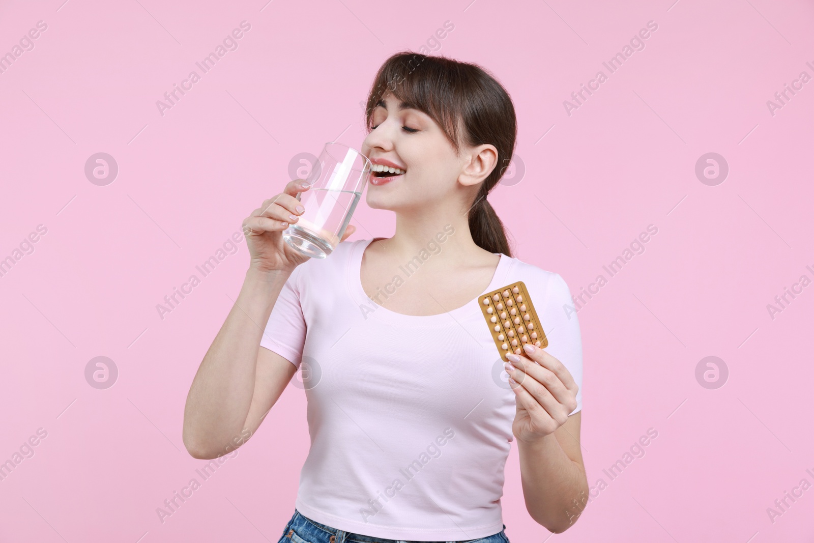 Photo of Young woman with contraceptive pills drinking water on pink background