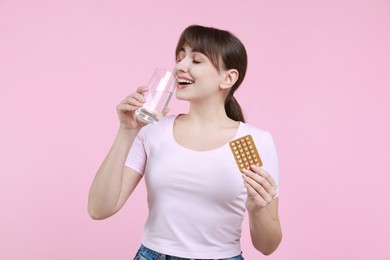 Young woman with contraceptive pills drinking water on pink background