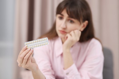 Upset woman with blister of contraceptive pills indoors, selective focus