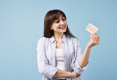 Photo of Smiling woman with blister of contraceptive pills on light blue background