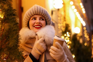 Happy woman with paper cup of coffee on city street, space for text. Christmas season