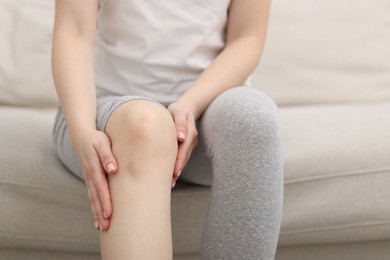 Photo of Young woman suffering from pain in knee on sofa at home, closeup