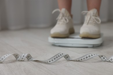 Photo of Woman on scales and measuring tape on floor indoors, selective focus. Weight loss