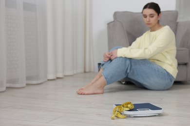 Photo of Depressed woman, scales and measuring tape on floor at home, selective focus with space for text. Bulimia problem