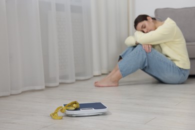 Photo of Depressed woman, scales and measuring tape on floor at home, selective focus with space for text. Bulimia problem