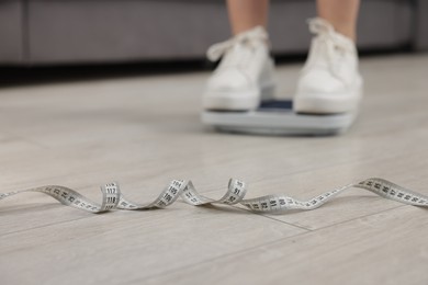 Photo of Woman on scales and measuring tape on floor indoors, selective focus. Weight loss