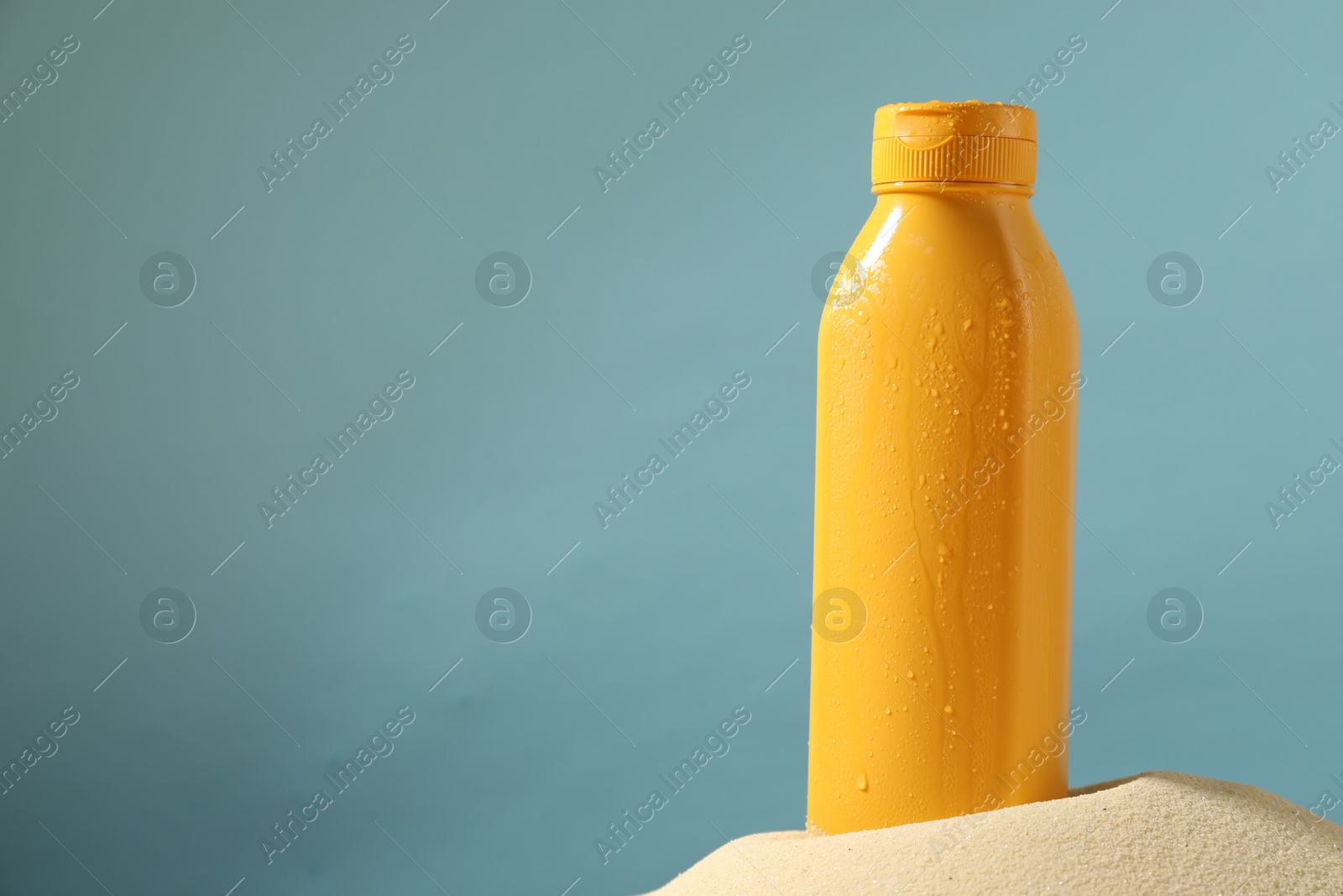 Photo of Bottle of sun protection product on sand against light blue background, space for text