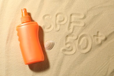 Photo of Abbreviation SPF 50 written on sand, bottle of sun protection product and seashell at beach, top view