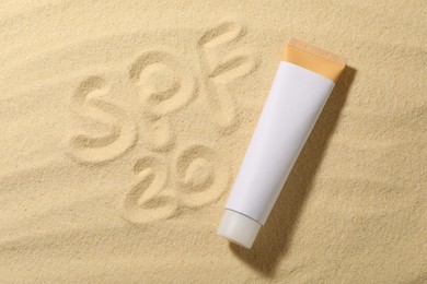 Photo of Abbreviation SPF 20 written on sand and tube of sun protection product at beach, top view