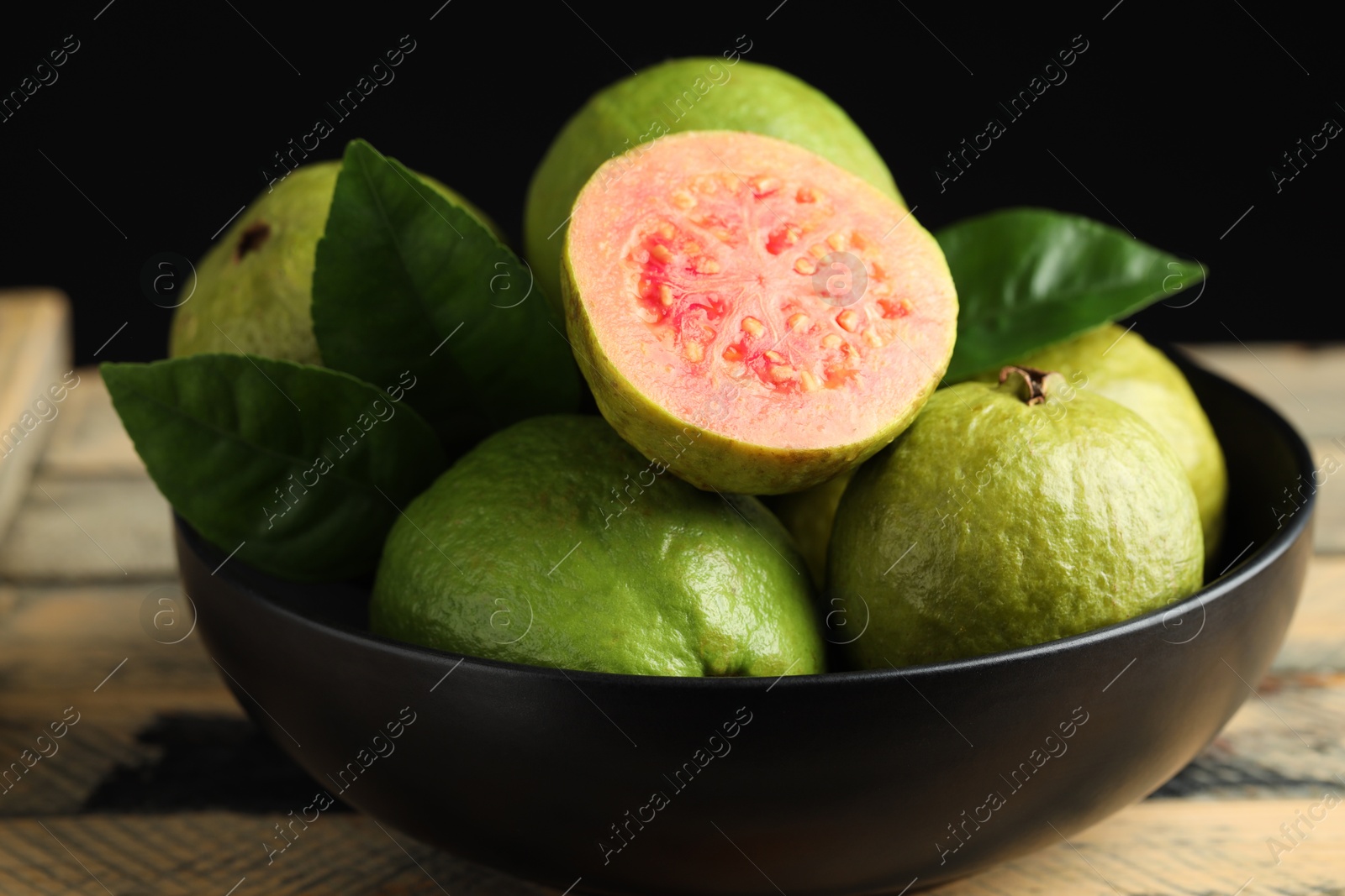 Photo of Fresh guava fruits in bowl on wooden table against black background, closeup