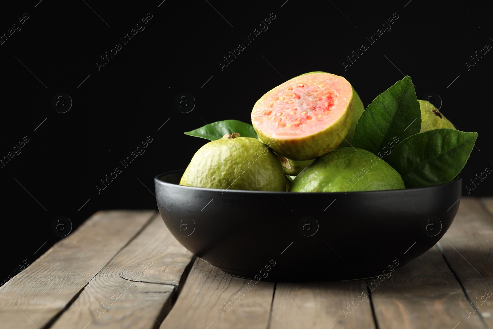 Photo of Fresh guava fruits in bowl on wooden table against black background, closeup