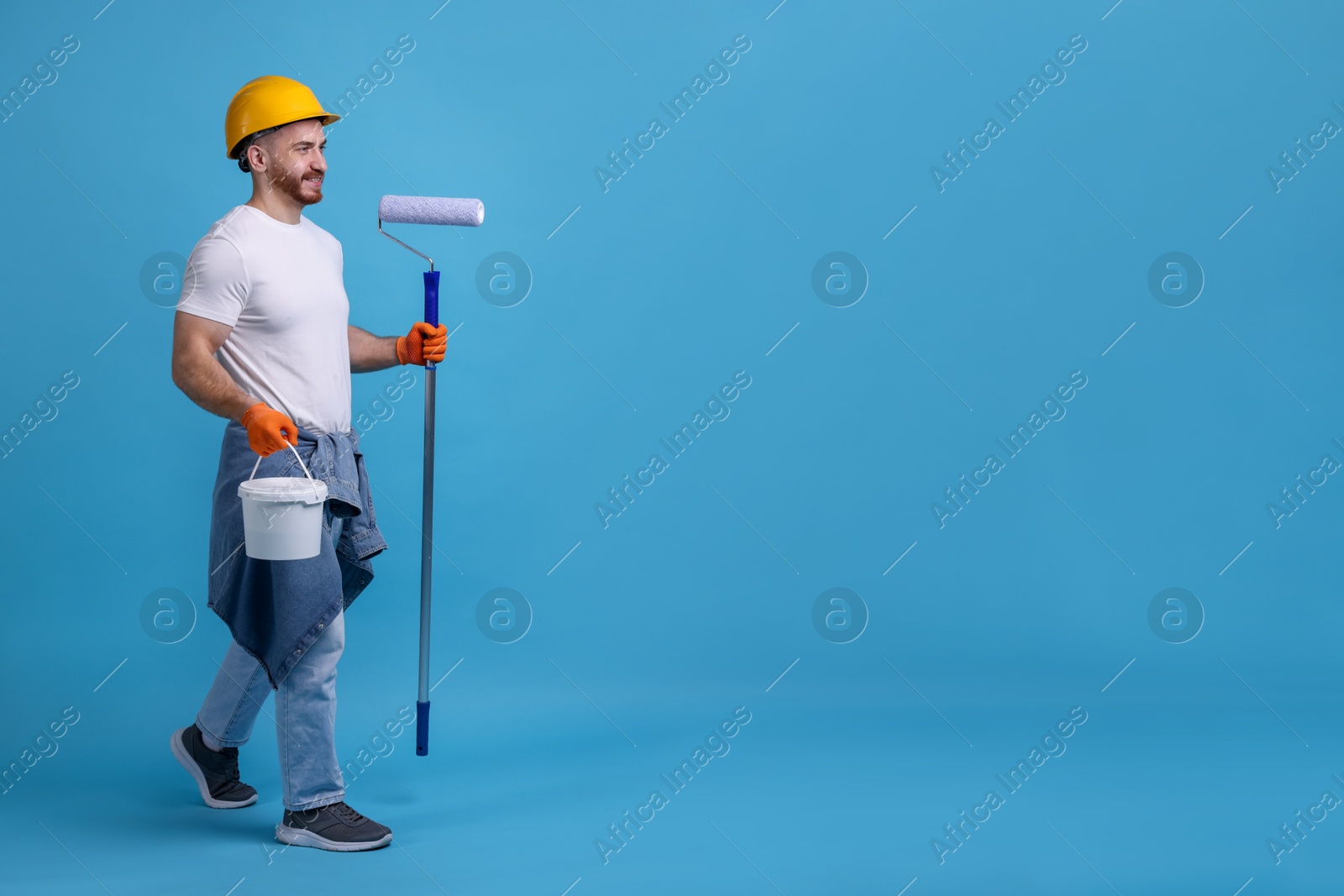 Photo of Man wearing hardhat with roller and bucket of paint on blue background. Space for text