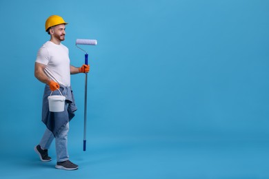 Man wearing hardhat with roller and bucket of paint on blue background. Space for text