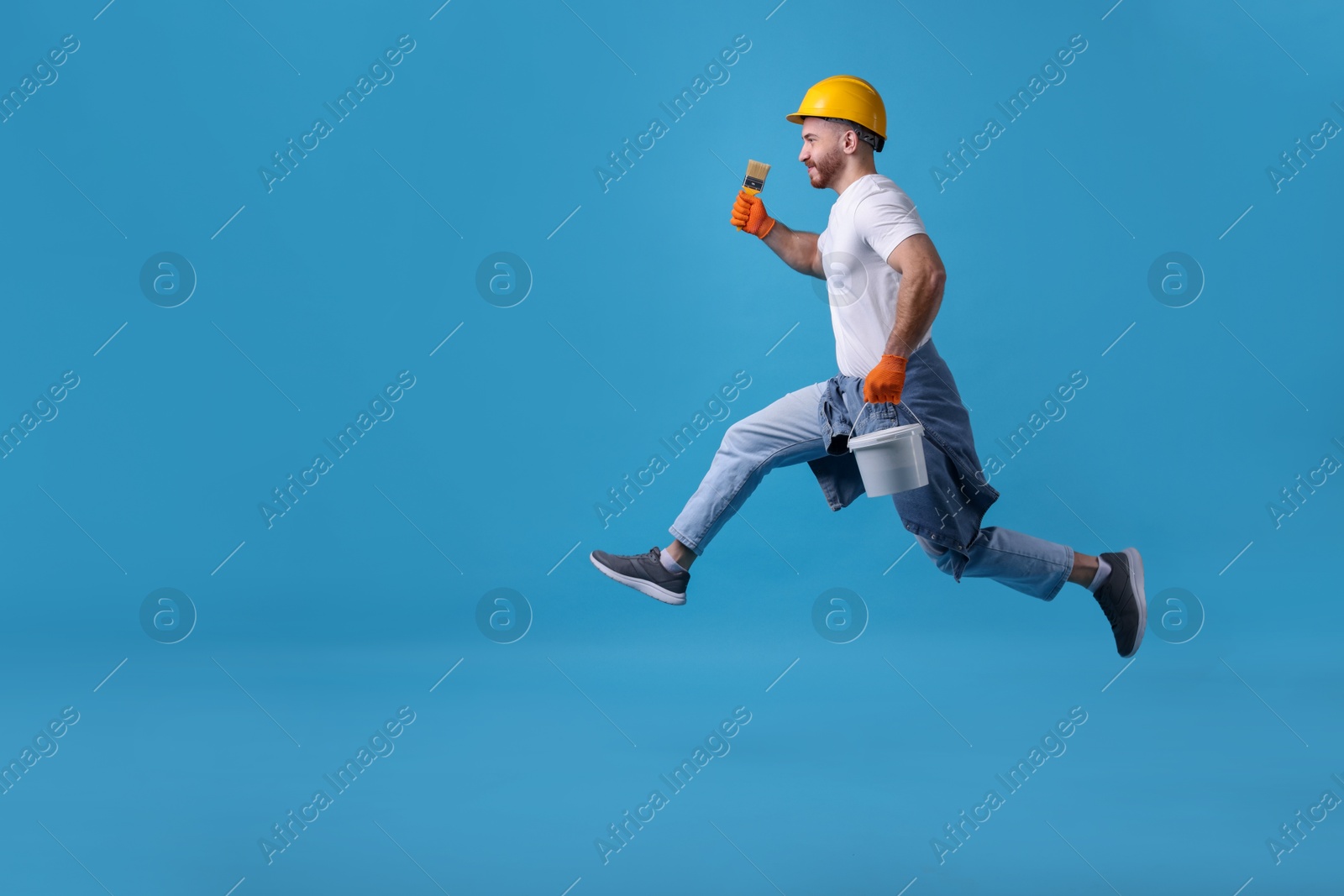 Photo of Man wearing hardhat with brush and bucket of paint jumping on blue background. Space for text