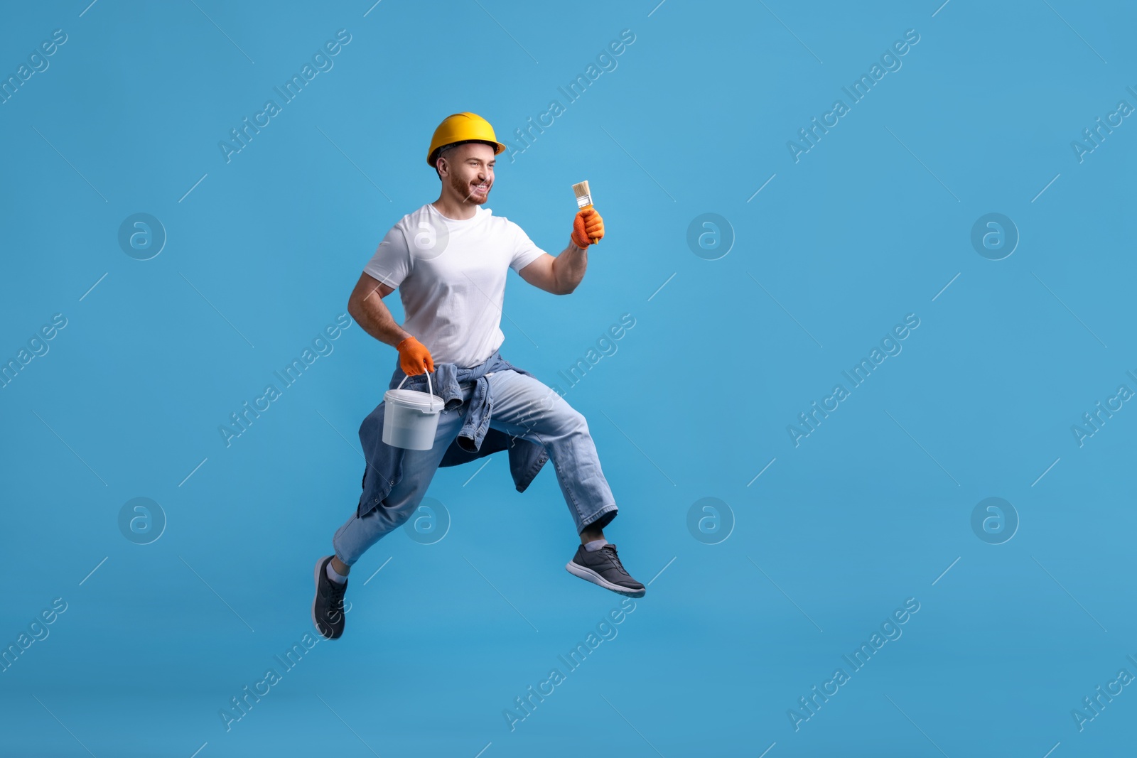 Photo of Man wearing hardhat with brush and bucket of paint jumping on blue background