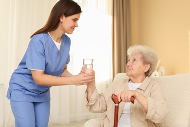 Photo of Healthcare worker giving elderly woman glass of water indoors