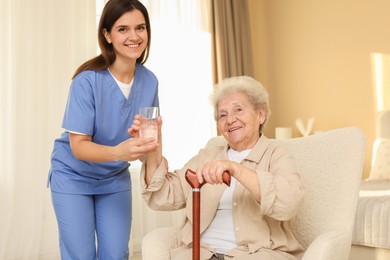 Photo of Healthcare worker giving elderly woman glass of water indoors