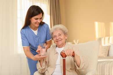 Photo of Healthcare worker giving elderly woman glass of water indoors