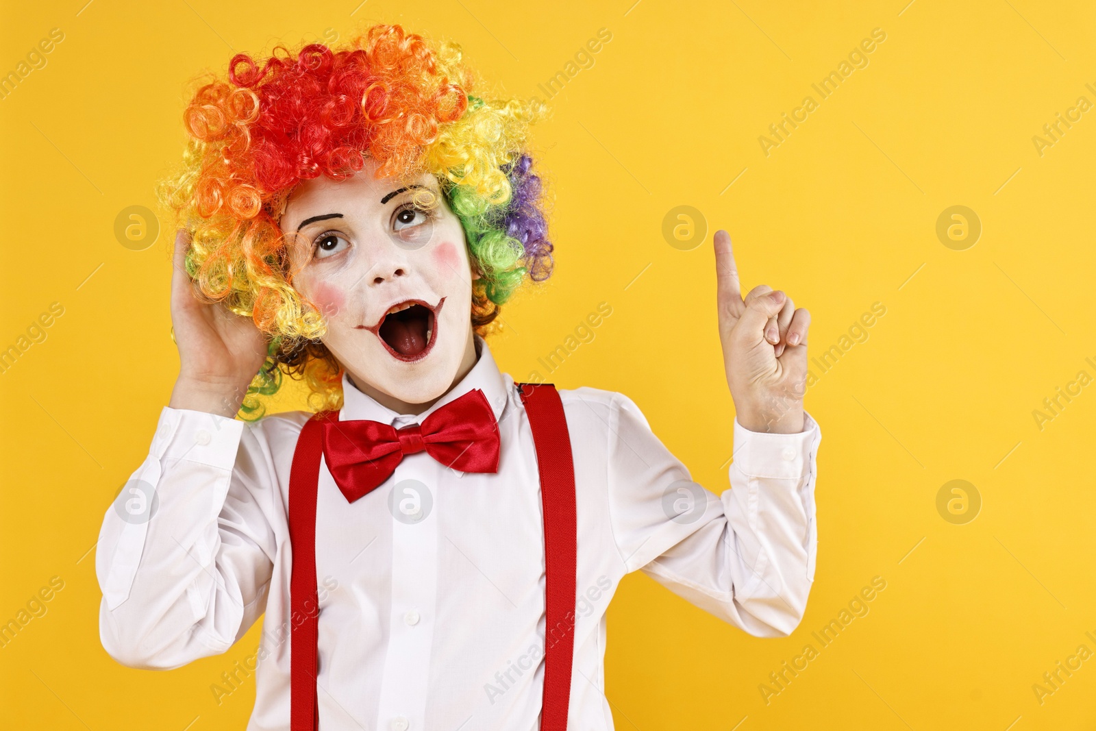 Photo of Emotional little boy dressed like clown pointing upwards on yellow background, space for text. Surprise party