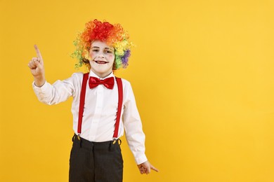 Photo of Emotional little boy dressed like clown pointing upwards on yellow background, space for text. Surprise party