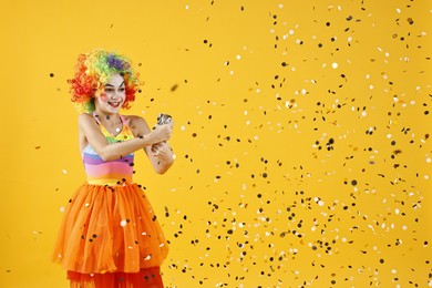 Photo of Happy girl dressed like clown with confetti popper on yellow background, space for text. Surprise party