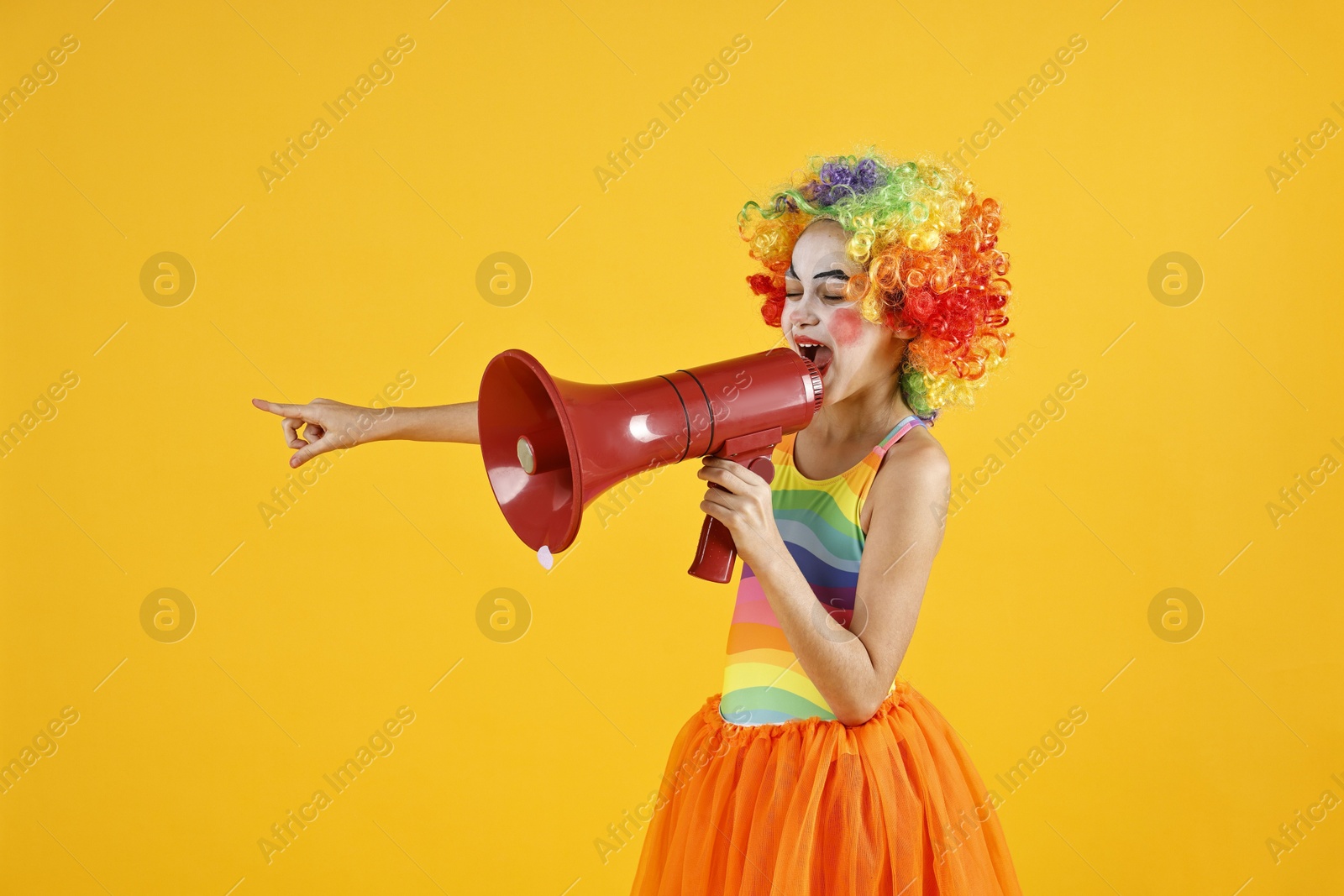 Photo of Cute girl dressed like clown shouting in megaphone and pointing at something on yellow background. Surprise party