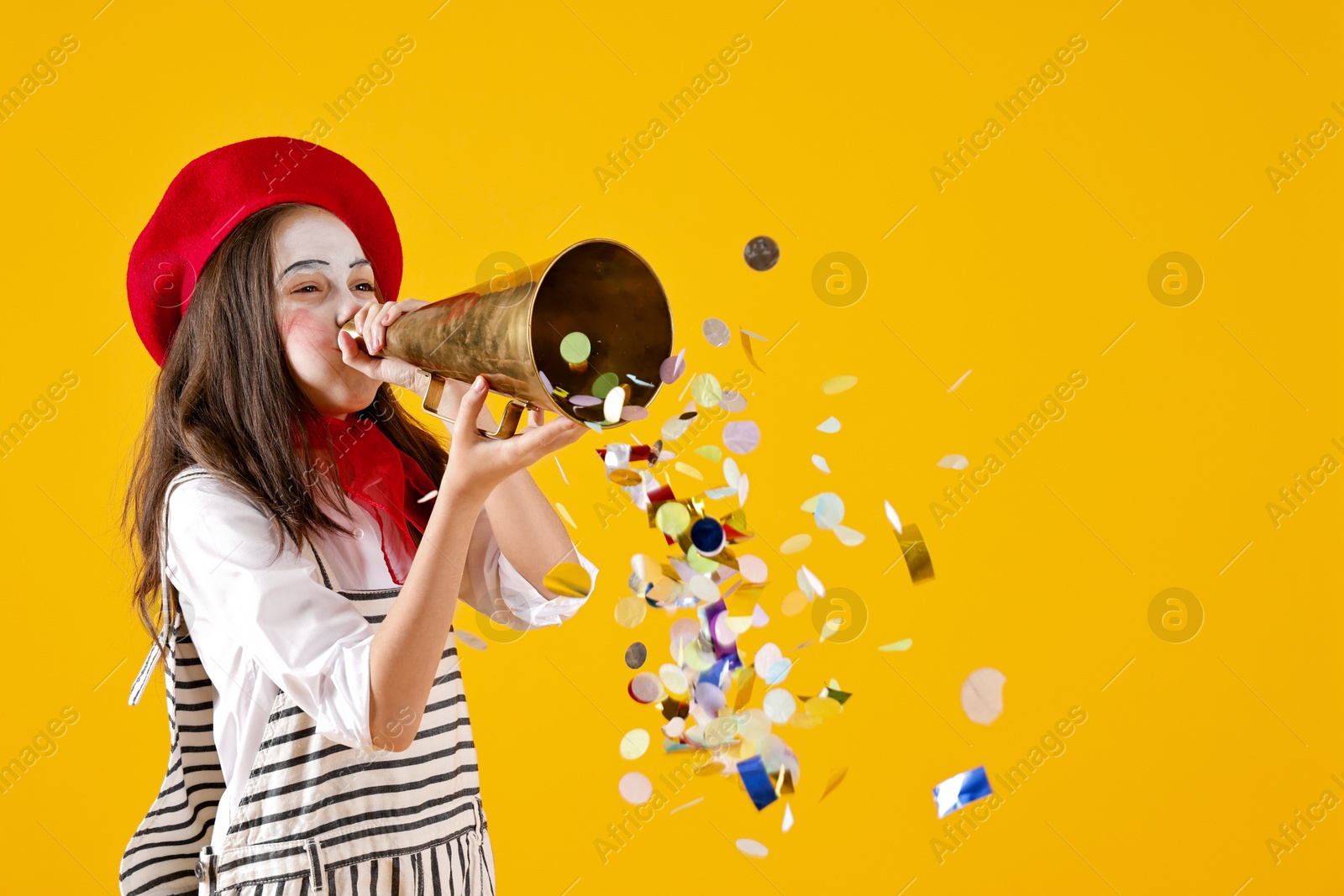 Photo of Happy girl dressed like mime with megaphone and confetti on yellow background, space for text. Surprise party