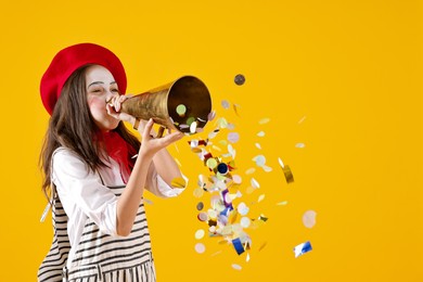 Happy girl dressed like mime with megaphone and confetti on yellow background, space for text. Surprise party