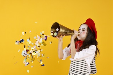 Happy girl dressed like mime with megaphone and confetti on yellow background. Surprise party