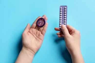 Woman holding blister of contraceptive pills and condom on light blue background, closeup