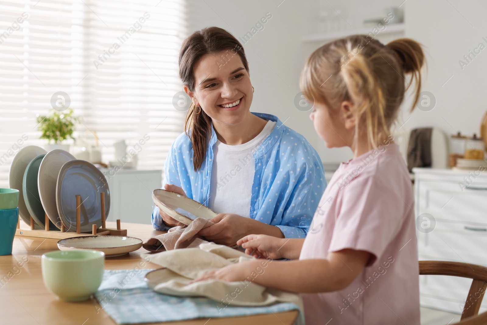 Photo of Little girl helping her mom wiping plates at table in kitchen