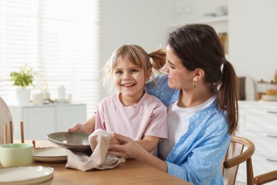 Photo of Little girl helping her mom wiping plates at table in kitchen