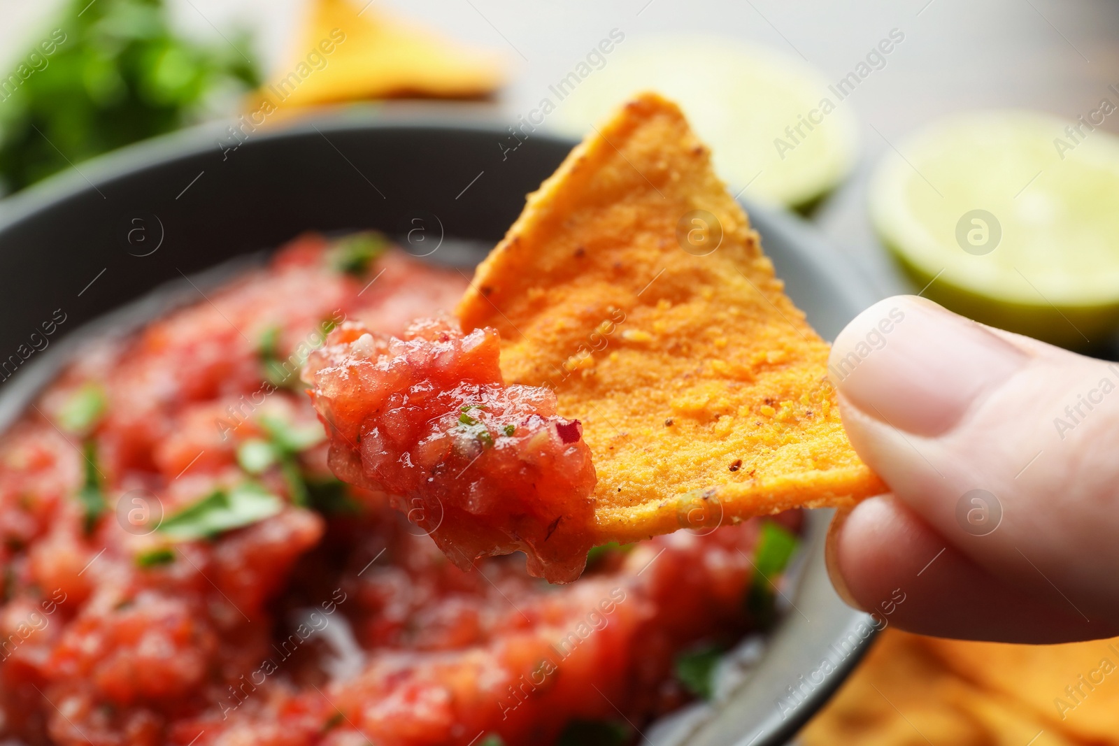 Photo of Woman holding nacho chip with spicy salsa sauce at table, closeup