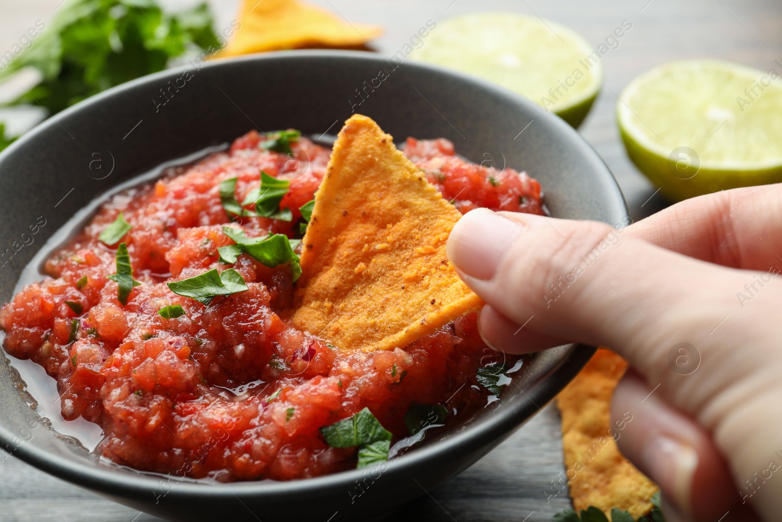 Photo of Woman dipping nacho chip into spicy salsa sauce at wooden table, closeup