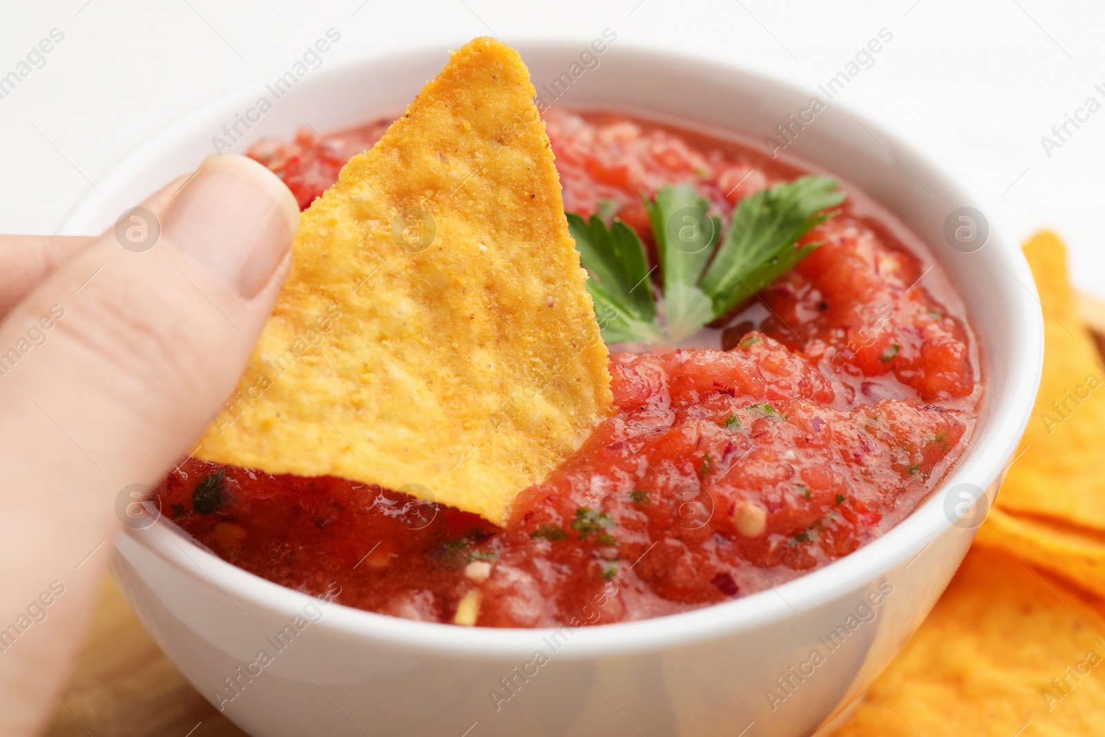 Photo of Woman dipping nacho chip into spicy salsa sauce at table, closeup