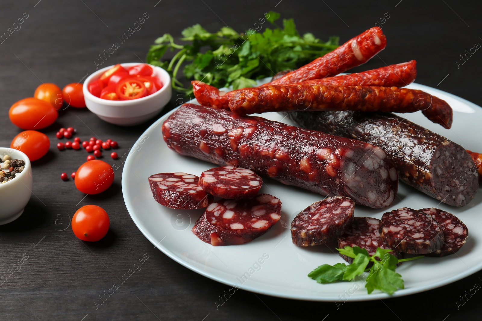 Photo of Different smoked sausages, cherry tomatoes and spices on dark wooden table, closeup