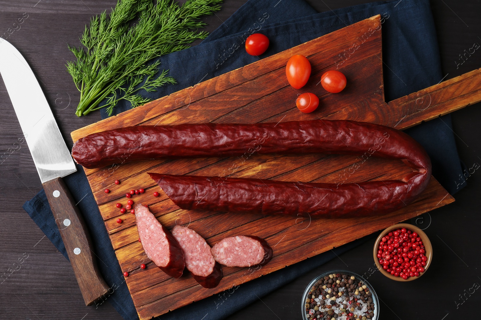 Photo of Delicious thin smoked sausage, cherry tomatoes, spices and knife on dark wooden table, flat lay