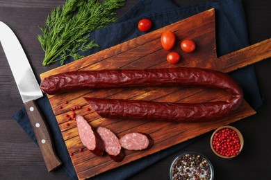 Photo of Delicious thin smoked sausage, cherry tomatoes, spices and knife on dark wooden table, flat lay