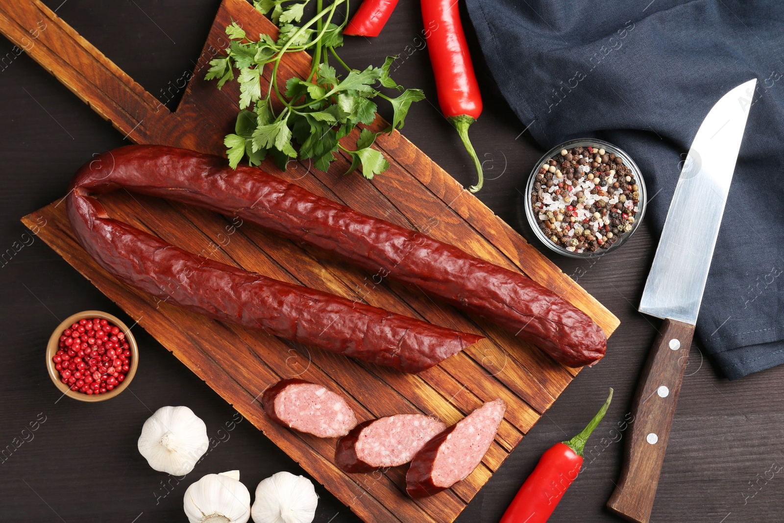 Photo of Delicious thin smoked sausage, spices and knife on dark wooden table, flat lay